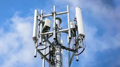 Cellular tower apex featuring a cluster of panel antennas and microwave dishes against a bright, cloudy sky.