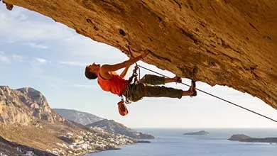 Rock climber on overhanging cliff, ocean and coastal village below.