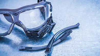 Safety goggles with a dark frame and elastic strap, shown with two detachable arms placed beside them on a textured blue surface.