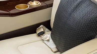 Close-up of a cream leather seat with a black woven pillow, a seatbelt with a gold buckle, and a dark wood console featuring two gold-rimmed cupholders.
