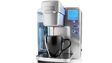 A silver single-serve coffee maker with a blue-lit display brews coffee into a black mug.