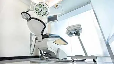 A black and white dental chair in a brightly lit exam room, with a large surgical light overhead.