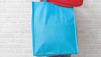 Person in a red shirt holding a large bright blue tote bag in front of a white brick wall.