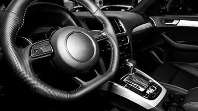 Black and white photo of a modern car interior from the driver’s view, showing a leather steering wheel with control buttons, dashboard gauges, center console with automatic gear shift, and driver’s side door panel.