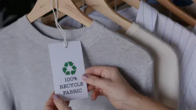 A person holds the tag of a gray sweater on a clothing rack. The tag displays a green recycling symbol and the text "100% RECYCLED FABRIC."