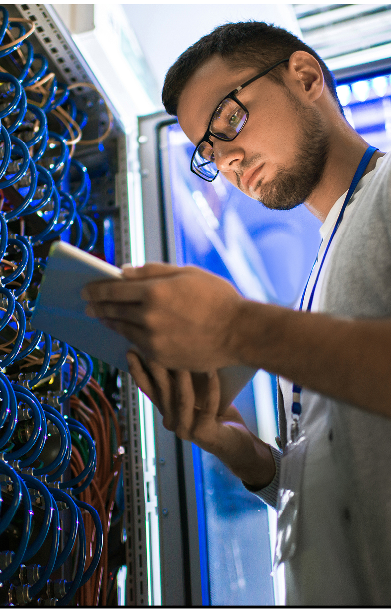 Low angle portrait of young man using digital tablet standing by server cabinet while working with supercomputer in blue light